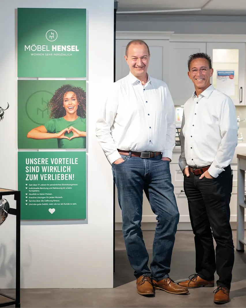 Zwei Männer stehen vor einer Küche, links grüne Poster mit Text Möbel Hensel und Unsere Vorteile sind wirklich zum Verlieben